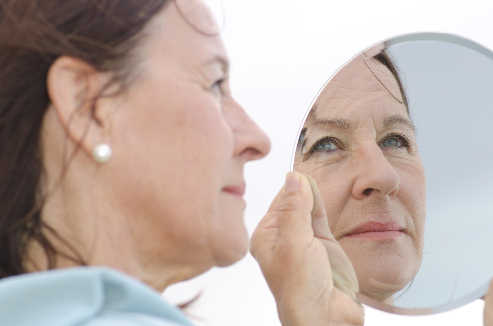 A woman holds a small, round mirror up to her face, gazing into it with a reflective expression for patient empowerment.
