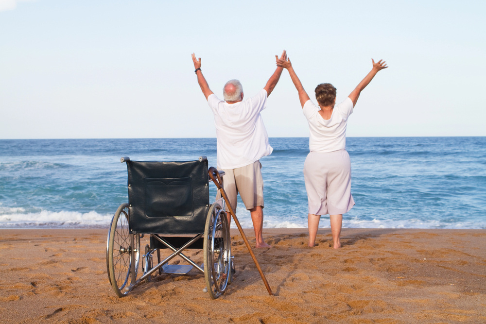 Two elderly individuals stand on a sandy beach facing the ocean, arms raised in joyful celebration.