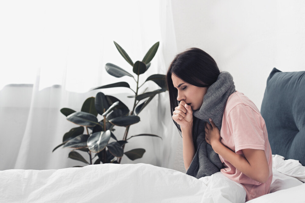 A woman wearing a thick grey scarf sits in bed and coughs into her hand while holding her chest.