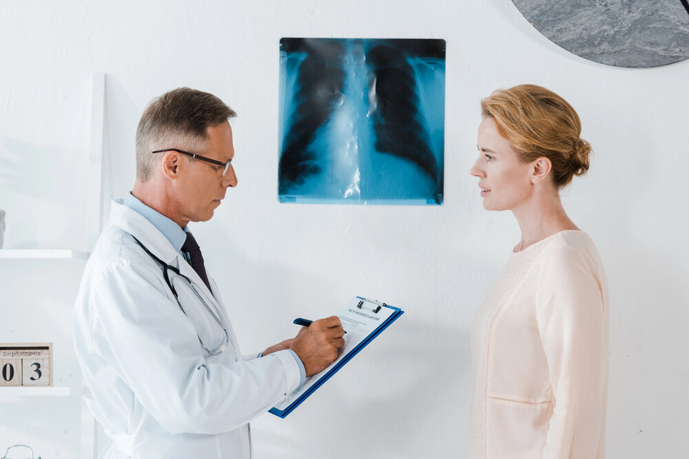A photograph of a male doctor in a white coat writing on a clipboard while standing across from a female patient, with a chest X-ray mounted on the wall behind them.