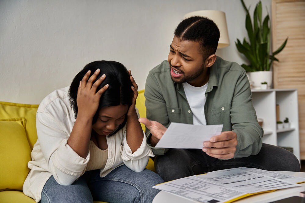 A man looks frustrated while holding a document and gesturing toward a woman who is sitting on a yellow sofa with her head in her hands.