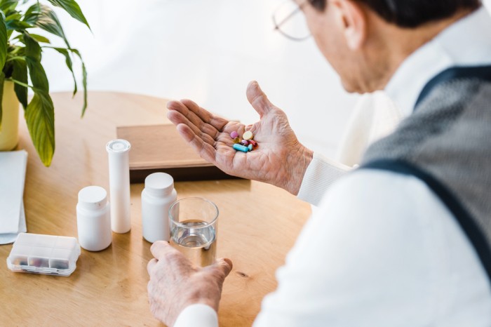 Older individual sitting at a table holding colorful pills and a glass of water, surrounded by pill bottles and a medication organizer to depict daily health management.