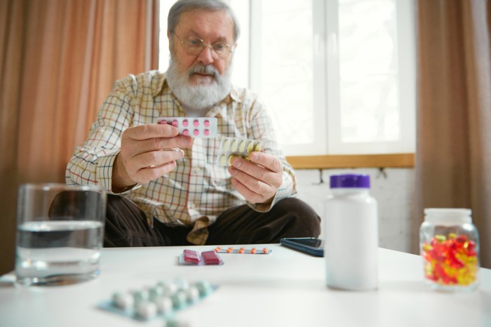 Older adult seated at a table organizing multiple blister packs of pills, capsules, and a pill bottle, highlighting medication management and daily healthcare routines.
