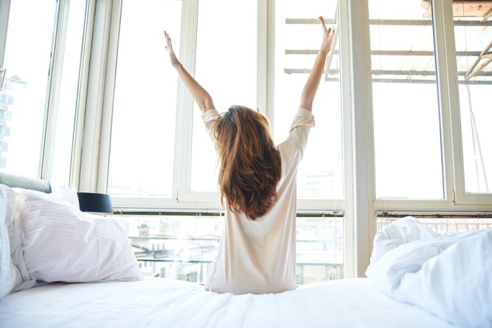 Woman stretching on bed in a bright bedroom with large windows in the morning
