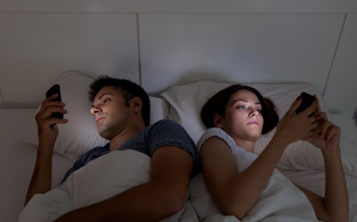 Couple lying in bed at night using smartphones, illuminated by screen light in a dark bedroom