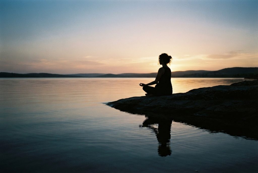 Person meditating on a rocky lakeshore at sunset with calm water and distant hills in the background.